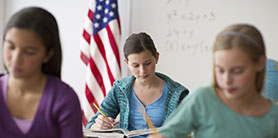 three students sitting and writing with an American flag behind them in a classroom
